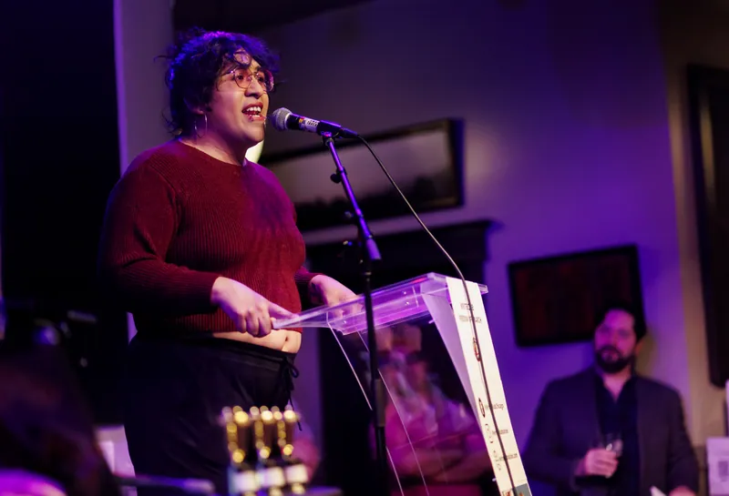 A combatant in a maroon sweater at the lectern, the crowd behind